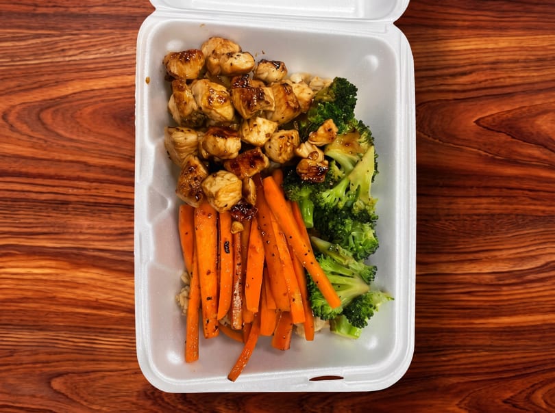 Takeout container with glazed chicken, steamed broccoli, and seasoned carrots on a wooden table.