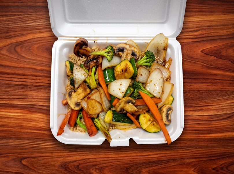 Open takeout container filled with stir-fried vegetables and rice on a wooden table.