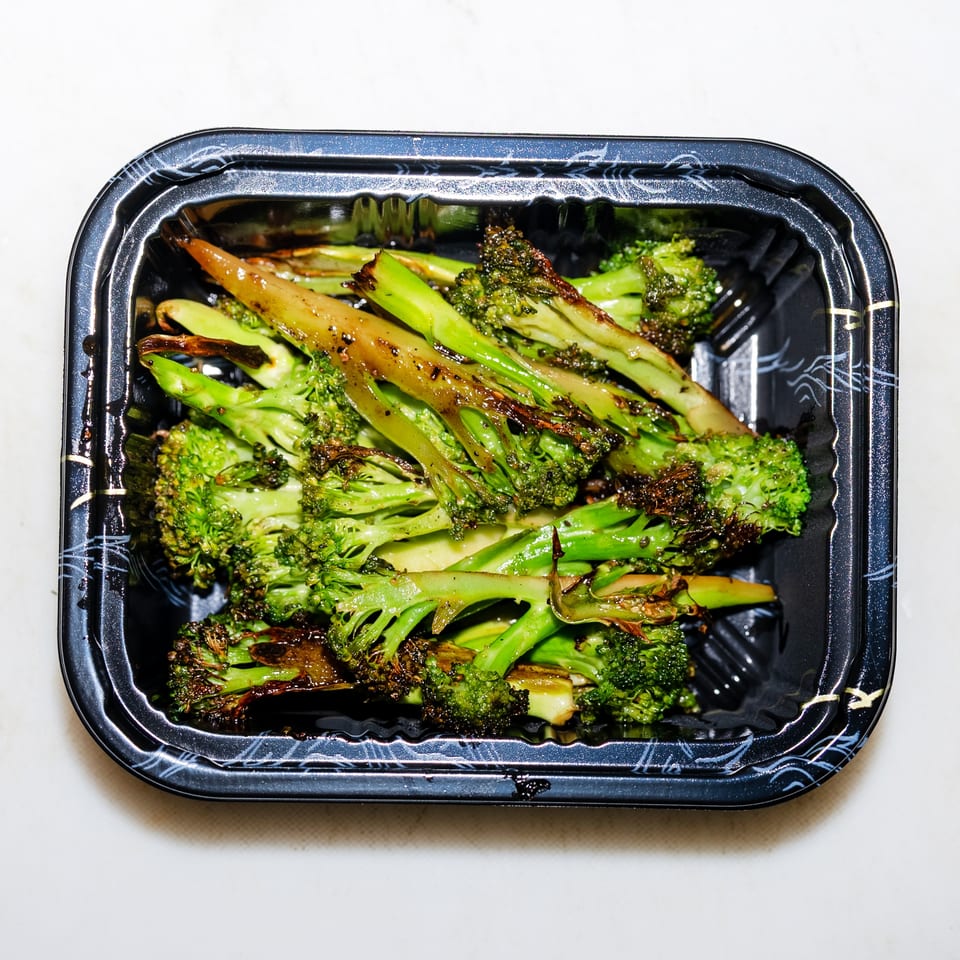 Roasted broccoli florets and stems glistening with seasoning in a black takeout container.