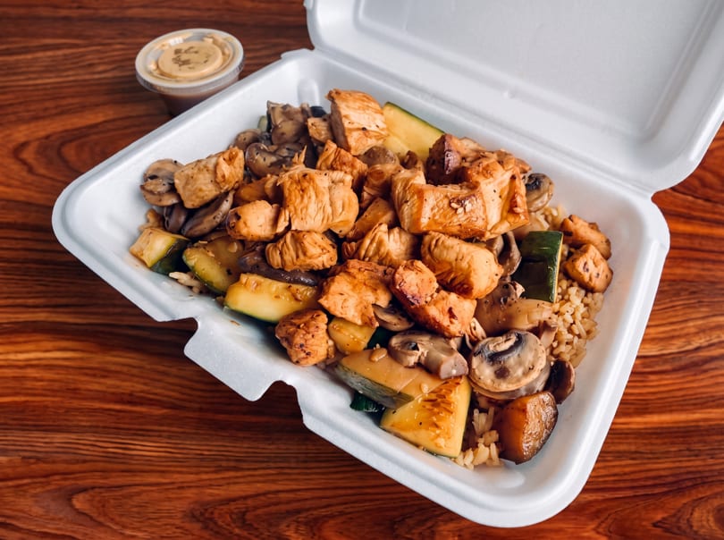 Takeout container with chicken, mushrooms, zucchini, and rice, plus a side of sauce on a wooden table.