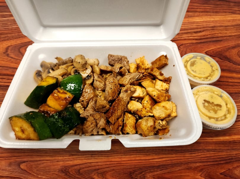 Open takeout container with chicken, beef, mushrooms, zucchini, and two sauce cups on a wooden table.