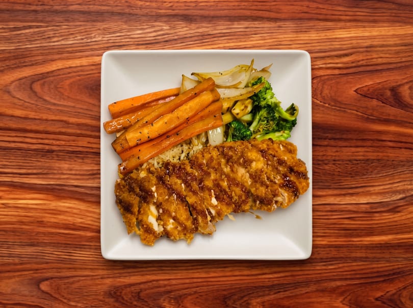 Crispy chicken katsu with rice, roasted carrots, broccoli, and onions on a white plate.