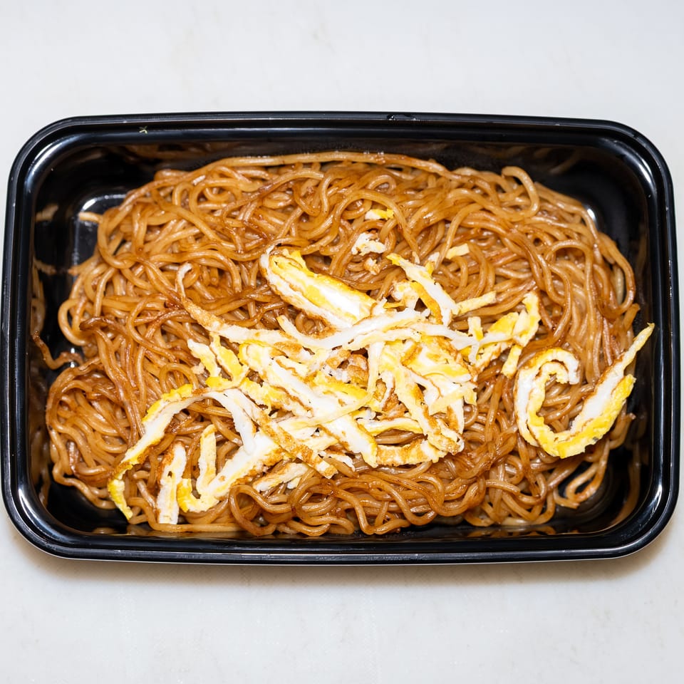 Soy-sauce noodles topped with shredded fried egg in a black rectangular takeout container.